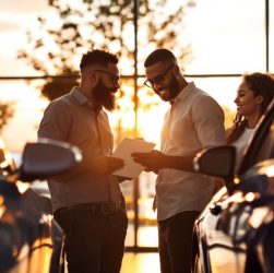Un couple examine et compare deux berlines neuves devant une concession automobile moderne au coucher du soleil, ouvrant les portes et regardant l'habitacle, voitures argent et bleu marine, lumière chaude et volumétrique.