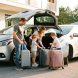 Un homme, une femme et deux enfants chargent des valises dans une voiture moderne devant une maison contemporaine, sous une lumière naturelle.
