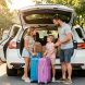 Une famille charge des valises dans une voiture familiale moderne avant un départ en vacances, sous une lumière naturelle.