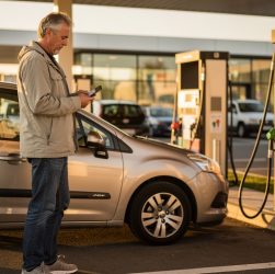 Un automobiliste consulte son smartphone devant une station-service française non identifiable, dans la lumière chaleureuse de la fin de journée.