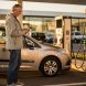 Un automobiliste consulte son smartphone devant une station-service française non identifiable, dans la lumière chaleureuse de la fin de journée.